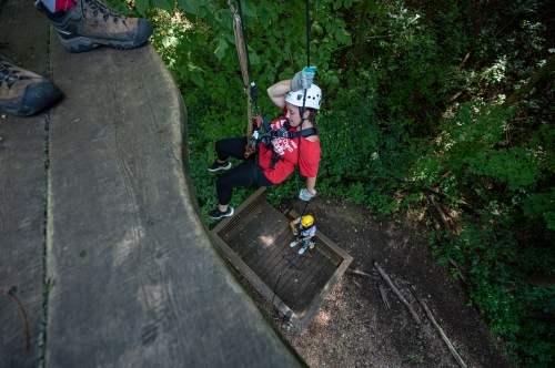 A person in a harness descends from a platform while another person waits below in a forested area.