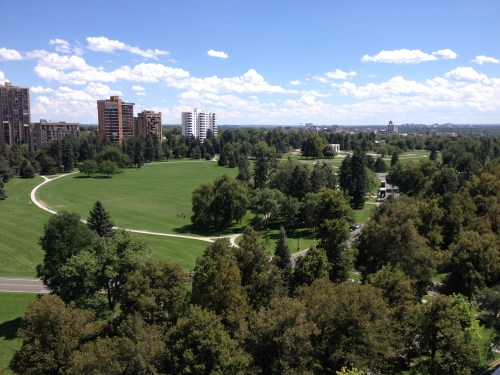 A panoramic view of a green park surrounded by trees and buildings under a blue sky with fluffy clouds.