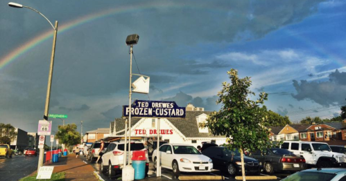 A colorful rainbow arcs over a frozen custard shop, with cars parked nearby under a cloudy sky.