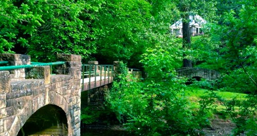 A stone bridge crosses a lush green area with trees, leading to a house in the background.