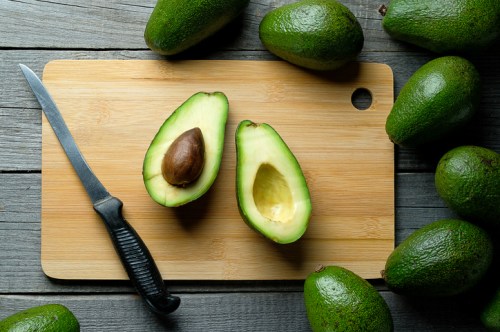 A halved avocado with a pit on a wooden cutting board, surrounded by whole avocados and a knife.
