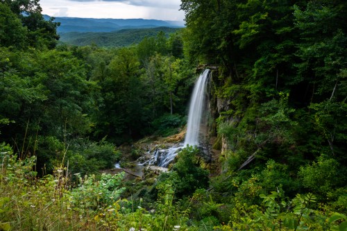 A serene waterfall cascades down rocky cliffs, surrounded by lush green trees and rolling hills in the background.