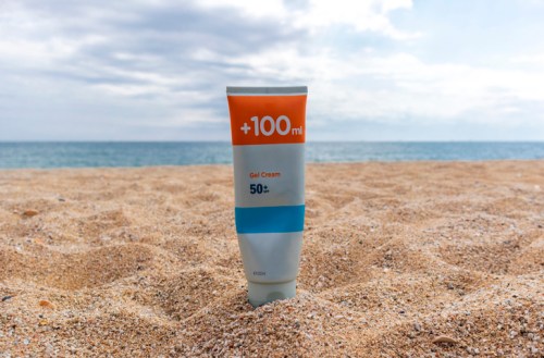 A tube of gel cream sunscreen with SPF 50 stands on sandy beach, with the ocean and cloudy sky in the background.