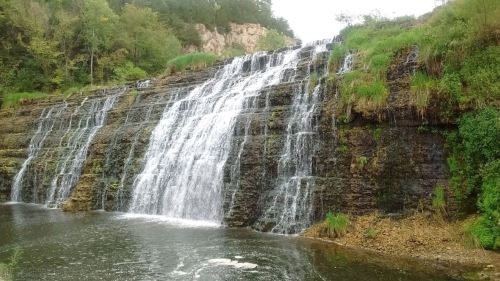A cascading waterfall flows over rocky cliffs into a serene pool, surrounded by lush greenery and trees.