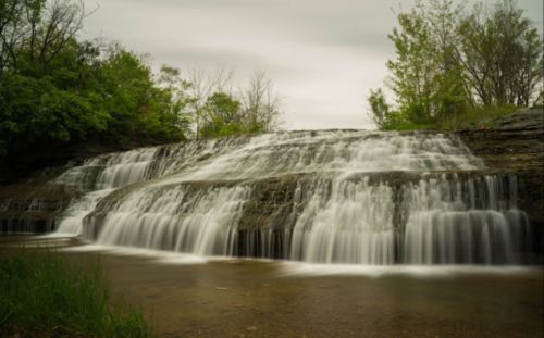 A serene waterfall cascading over rocky ledges, surrounded by lush greenery under a cloudy sky.
