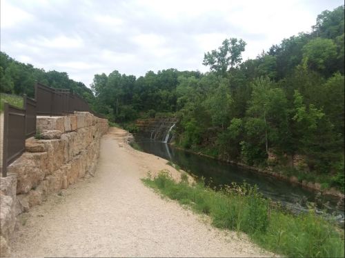 A scenic pathway alongside a river, with lush greenery and a small waterfall in the background under a cloudy sky.