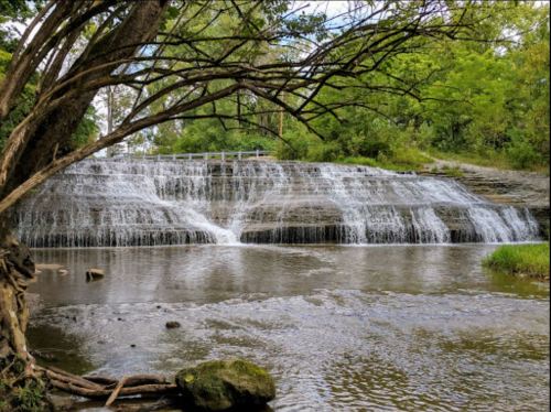 A serene waterfall cascades over rocky ledges, surrounded by lush greenery and a calm river.