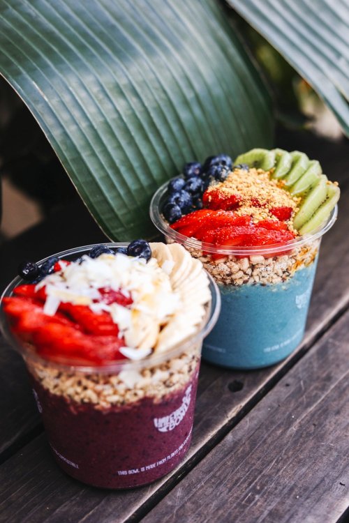 Two colorful smoothie bowls topped with fruits, granola, and coconut, placed on a wooden table with green leaves in the background.