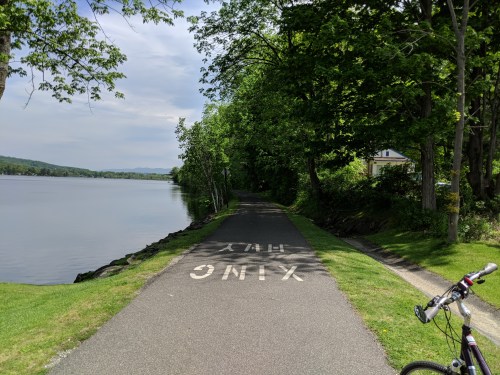 A scenic path along a lake, lined with trees, with a bicycle parked nearby and the word "XING" painted on the pavement.