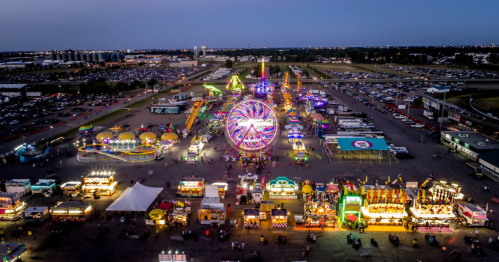Aerial view of a vibrant carnival at dusk, featuring rides, games, and colorful lights amidst a large parking area.