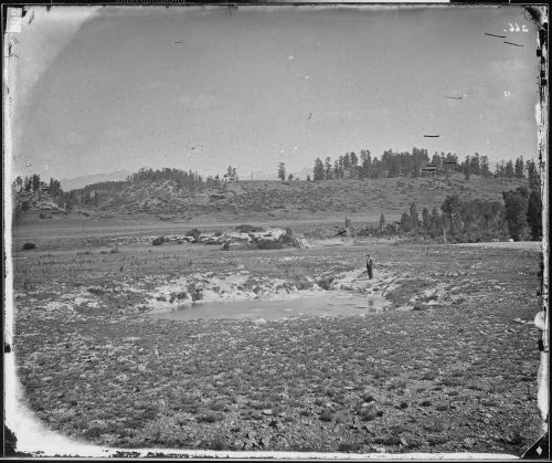A black-and-white landscape featuring a man standing by a small pond, surrounded by grassy fields and distant trees.