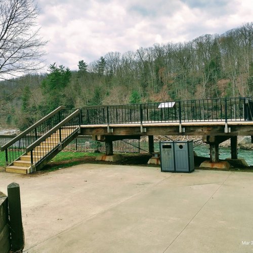 A wooden bridge over a river, with stairs leading up to it and a trash can nearby, surrounded by trees and hills.