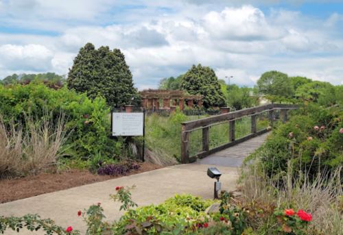 A wooden bridge leads through lush greenery and flowers, with a sign visible in a serene garden setting.