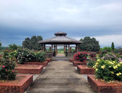 A gazebo surrounded by colorful rose bushes under a cloudy sky, with a pathway leading through the garden.