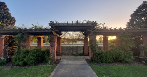 A brick pergola covered in greenery, leading to a pathway with a view of a sunset in the background.