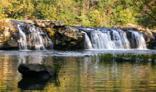 A serene waterfall cascades over rocks into a calm river, surrounded by lush greenery and trees.