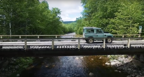 A light blue vehicle crosses a wooden bridge over a clear stream, surrounded by lush green trees.