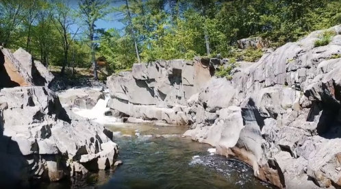 A serene river flows through rocky terrain, surrounded by lush greenery and clear blue skies.