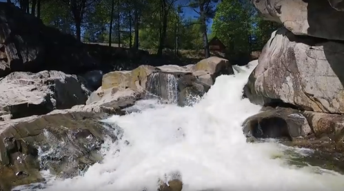 A cascading waterfall flows over rocky terrain, surrounded by lush greenery and a clear blue sky.
