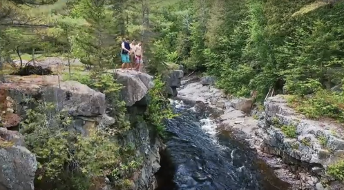 Two people stand on a rocky ledge overlooking a flowing river surrounded by lush greenery.