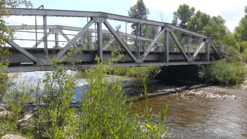 A metal bridge spans a calm river, surrounded by greenery and trees under a clear blue sky.