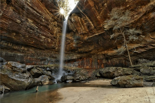 A serene waterfall cascades into a rocky pool, surrounded by towering cliffs and trees in a tranquil natural setting.