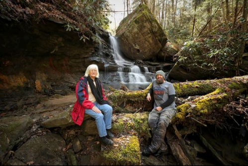 Two people sit on a mossy rock by a waterfall in a lush forest, surrounded by greenery and rocky terrain.
