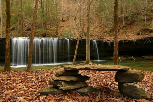 A serene forest scene featuring a small stone bench in front of a waterfall, surrounded by autumn leaves and trees.