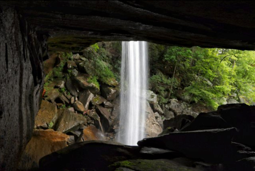 A waterfall cascades down rocks, framed by a cave entrance and surrounded by lush green foliage.