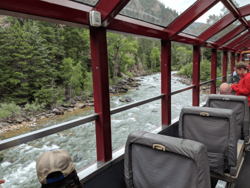 View from a train window showing a river and lush greenery along the banks, with passengers seated inside.