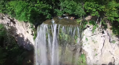 Aerial view of a cascading waterfall surrounded by lush green trees and rocky cliffs.