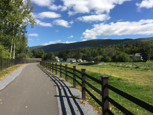 A scenic pathway lined with a wooden fence, surrounded by green fields and mountains under a blue sky with clouds.