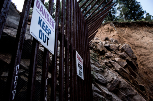 A rusted fence with "Danger Keep Out" signs near a steep, eroded cliffside in a wooded area.