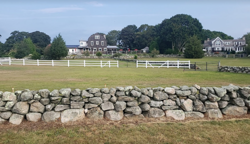 A scenic view of a grassy field with a stone wall, surrounded by trees and houses in the background.