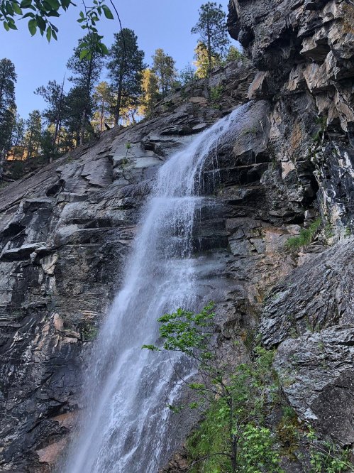 A cascading waterfall flows down a rocky cliff, surrounded by trees and greenery under a clear blue sky.