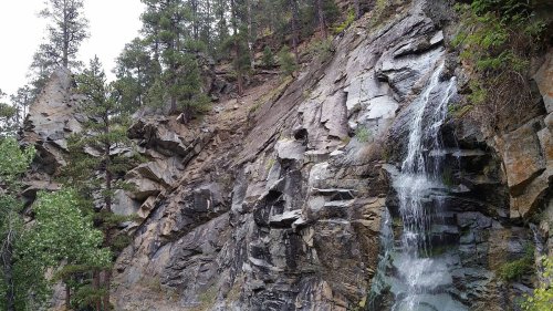 A cascading waterfall flows down rocky cliffs, surrounded by lush green trees and rugged terrain.