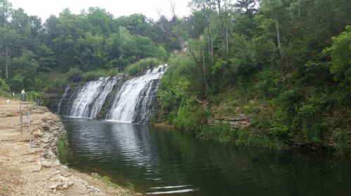 A serene waterfall cascades over rocks into a calm, green-tinted pool, surrounded by lush trees and foliage.