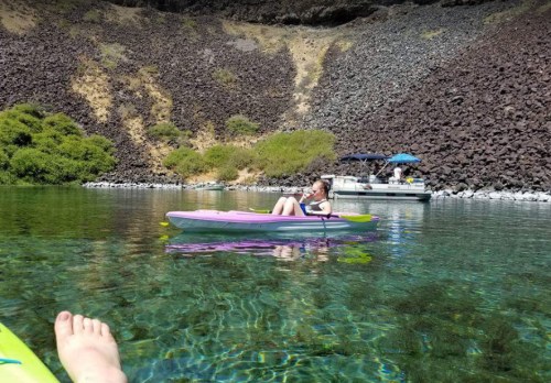 A person relaxes in a kayak on clear water, with a boat in the background and rocky cliffs nearby.