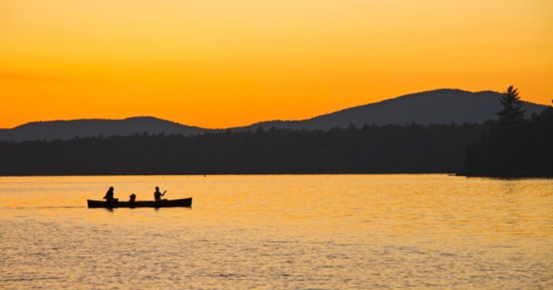 Two people canoeing on a calm lake at sunset, with silhouettes of mountains in the background.