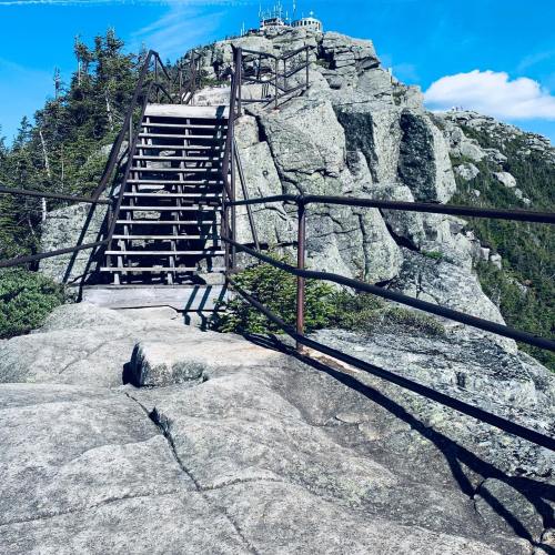 A metal staircase leads up a rocky mountain, surrounded by trees and a clear blue sky.