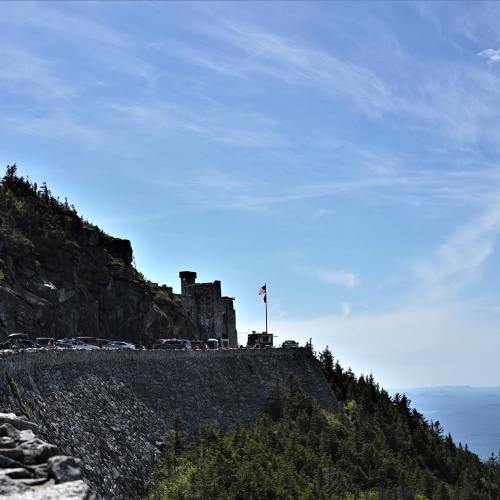 A scenic view of a rocky cliff with a flag, overlooking a parking area and lush greenery under a clear blue sky.
