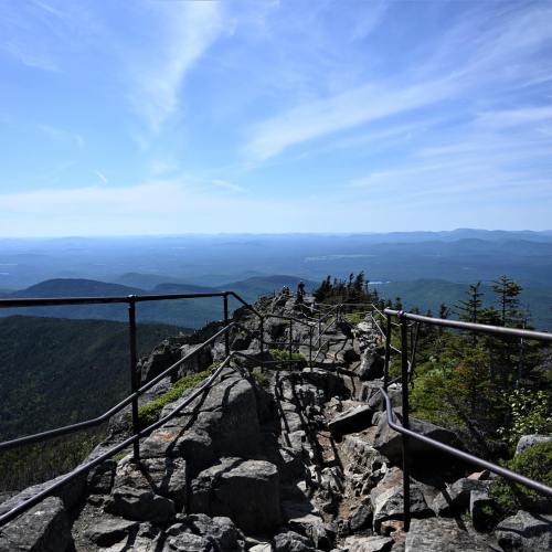 A rocky mountain trail with railings, overlooking a vast landscape of mountains and blue sky.
