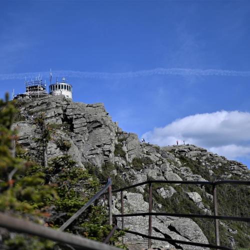 A rocky mountain peak with a lookout tower, under a blue sky with scattered clouds and a few hikers in the distance.