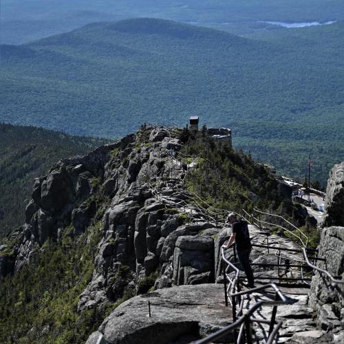 A person stands on a rocky ledge overlooking a vast green landscape and distant mountains under a clear blue sky.