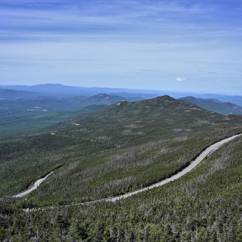A winding road cuts through lush green mountains under a clear blue sky, showcasing a scenic landscape.