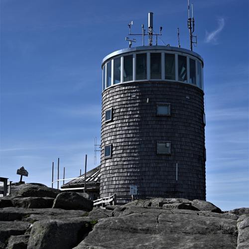 A tall, round wooden tower with a glass top, surrounded by rocky terrain and a clear blue sky.