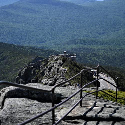 A scenic view from a rocky overlook, showing lush green mountains and a winding road below.