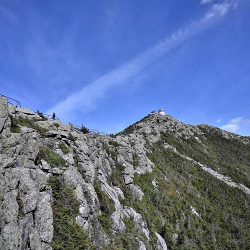A rocky mountain landscape under a blue sky, with a lookout tower at the summit and a trail leading up.