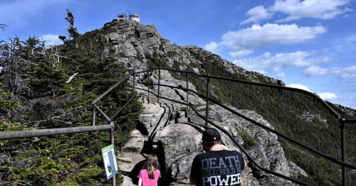 A person and a child walk along a rocky path towards a mountain summit with a lookout tower under a blue sky.
