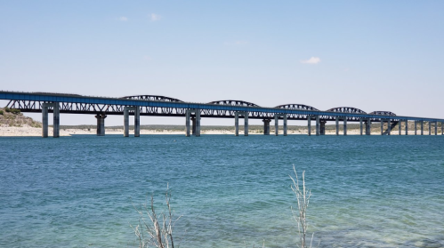 A long bridge spans across a clear blue lake under a bright sky, with gentle waves lapping at the shore.